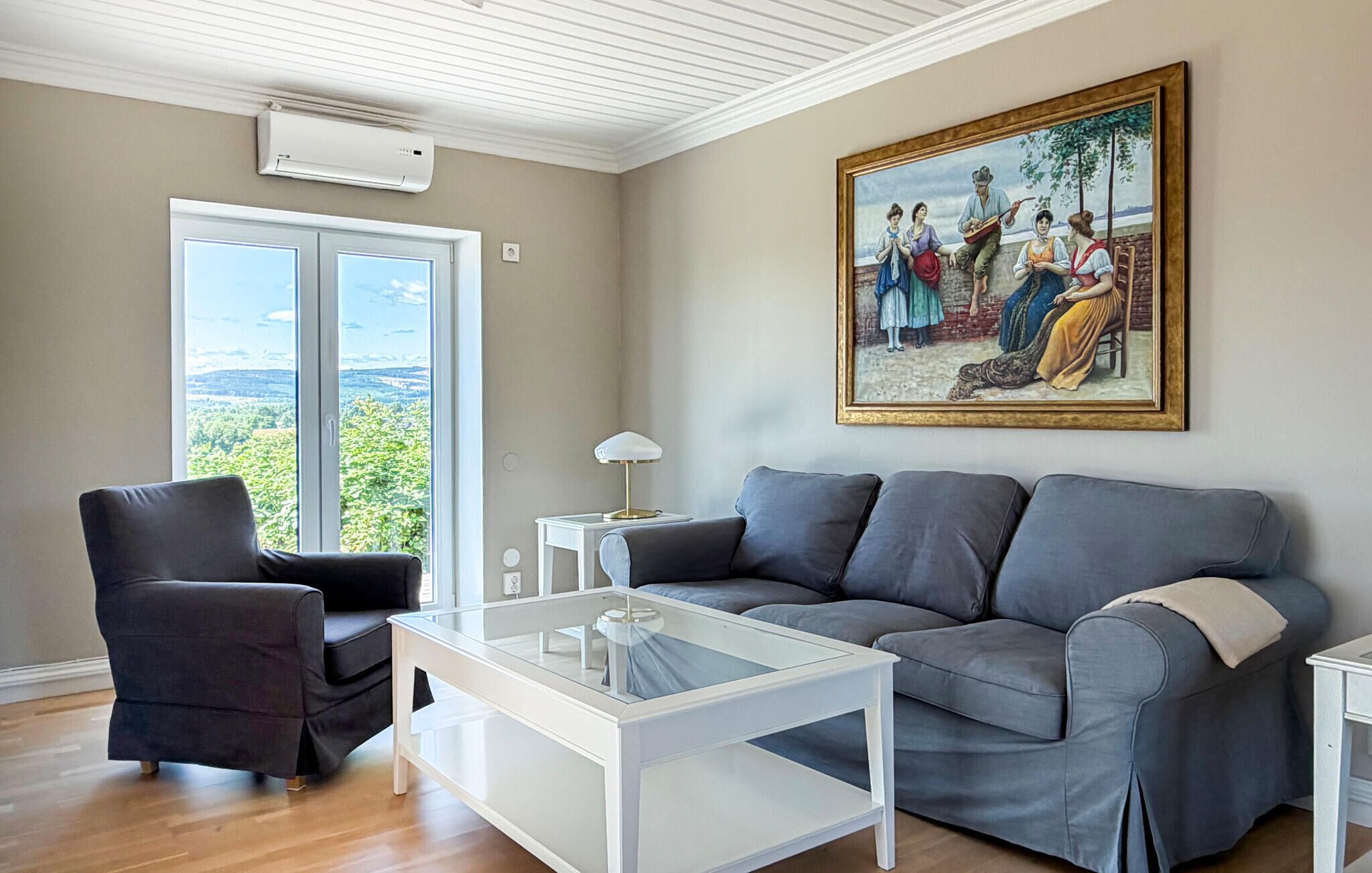 Ett vardagsrum med en grå soffa, ett soffbord i glas, en svart fåtölj, en tavla på väggen och ett stort fönster med utsikt.