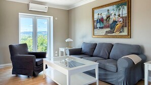 Una sala de estar con un sofá gris, una mesa de centro de vidrio, un sillón negro, un cuadro en la pared y un ventanal con vistas.