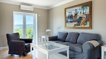 Ett vardagsrum med en grå soffa, ett soffbord i glas, en svart fåtölj, en tavla på väggen och ett stort fönster med utsikt.