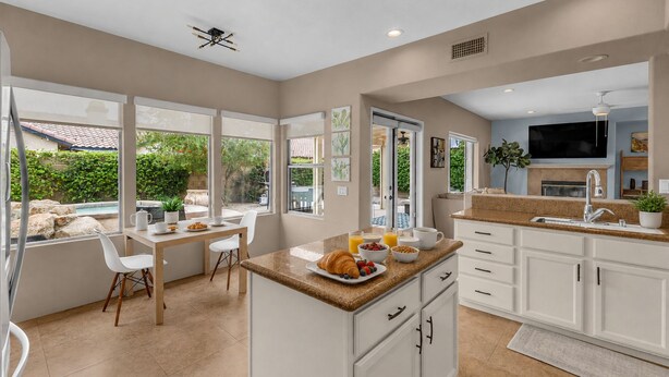 A modern kitchen with white cabinetry, a breakfast bar, and a dining area with a view of the outdoors. - Desert Sanctuary | Pool, Spa & Sleeps 12 (Indio)