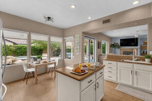 A modern kitchen with white cabinetry, a breakfast bar, and a dining area with a view of the outdoors. - Desert Sanctuary | Pool, Spa & Sleeps 12 (Indio)