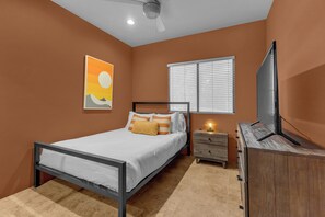 Ein Schlafzimmer mit Bett, Nachttisch, Kommode, Deckenventilator, Fenster mit Jalousien und einem an der Wand montierten Fernseher.