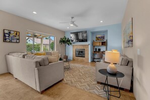 A living room with a sofa, a table with a lamp, a fireplace, and a ceiling fan. - Desert Sanctuary | Pool, Spa & Sleeps 12 (Indio)