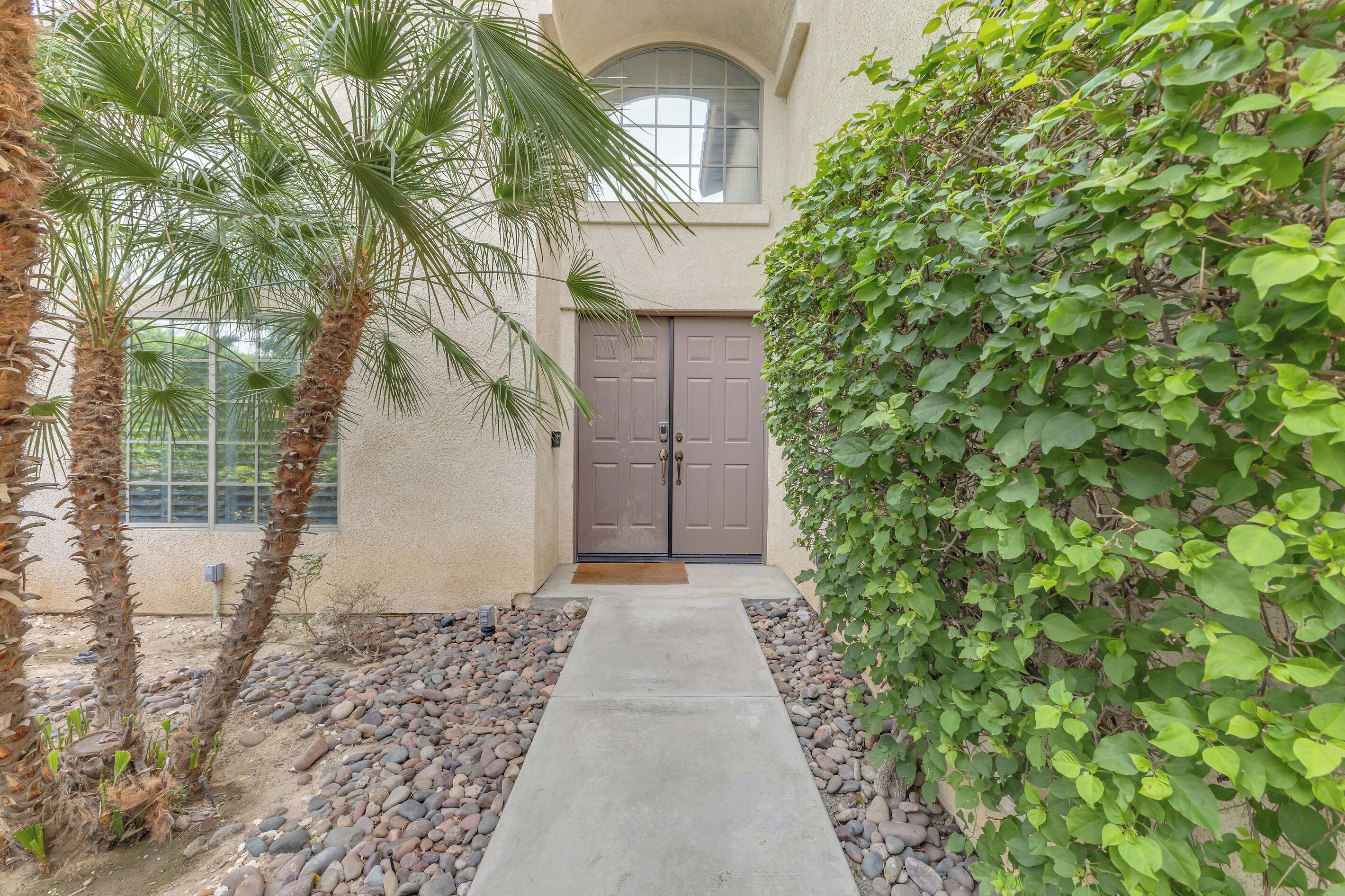 A pathway leading to a door with a window above, surrounded by palm trees and a green hedge.