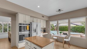 A modern kitchen with white cabinets, a granite countertop, and a dining area with a table and chairs. - Desert Sanctuary | Pool, Spa & Sleeps 12 (Indio)