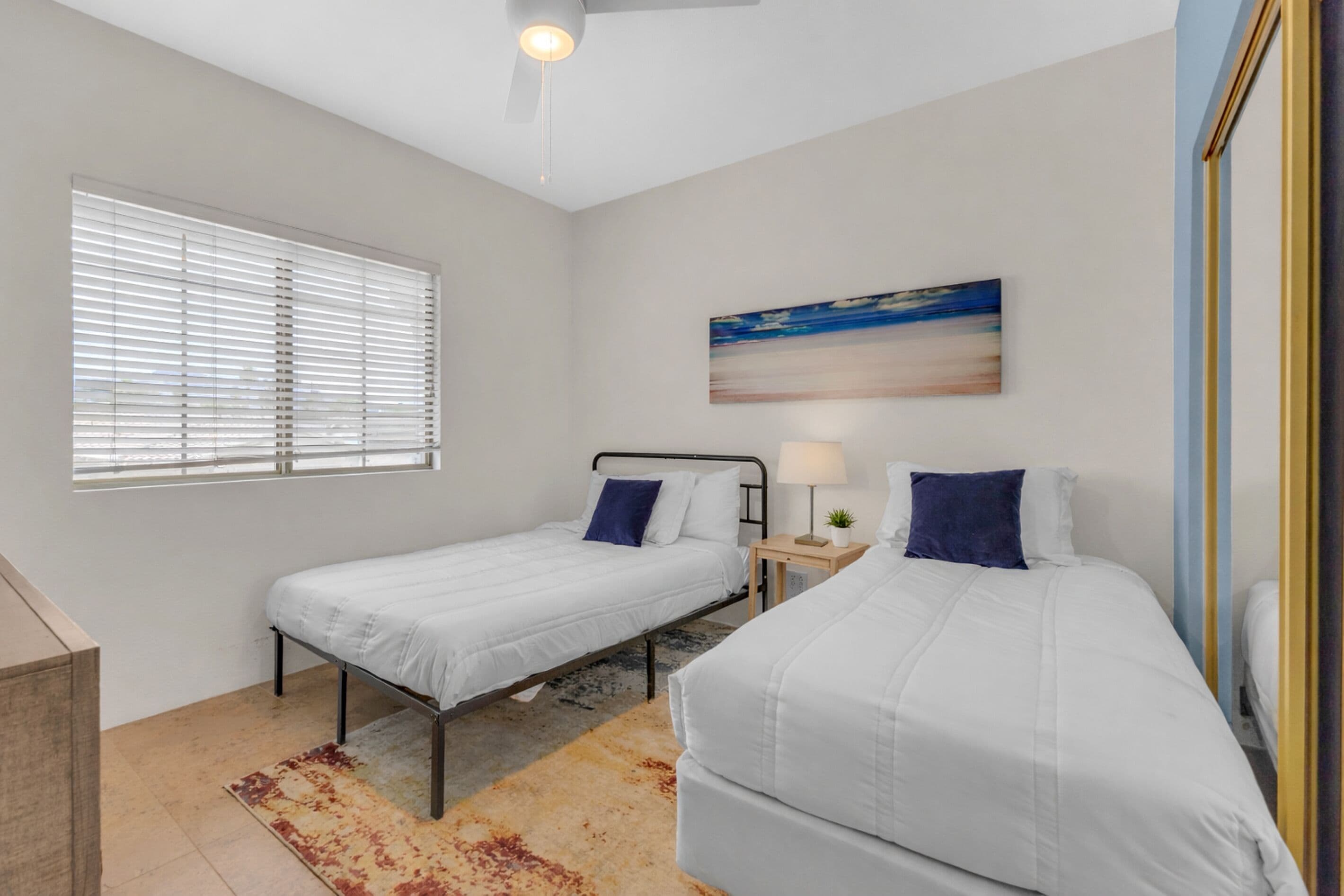 A bedroom with two single beds, a ceiling fan, a window with blinds, a painting on the wall, and a small bedside table with a lamp.