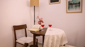 Ein Zimmer mit Holzstuhl, einem kleinen Tisch mit weißer Tischdecke, zwei Gläsern Rotwein, einem Buch und einer Lampe.