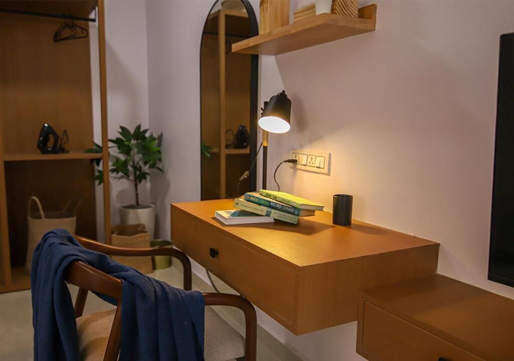 A wooden desk with a lamp, books, and a plant in a room with a wooden shelf and a hanging coat.