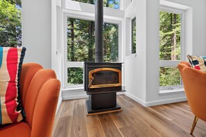 Ein Zimmer mit Kaminofen, großen Fenstern und einem modernen orangefarbenen Sessel.