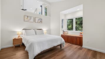 A bedroom with a bed, bedside tables, a window with a view of greenery, and artwork on the wall.