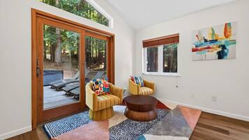A modern living room with a large sliding glass door leading to a deck, two armchairs with colorful cushions, a round wooden coffee table, and a painting on the wall.