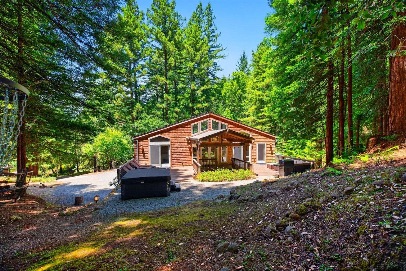 A cabin with a covered porch, a hot tub, and a gravel driveway surrounded by dense forest.