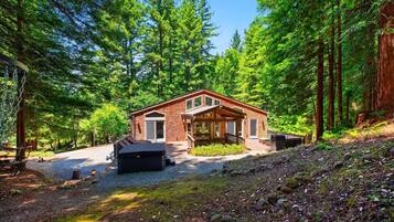 A cabin with a covered porch, a hot tub, and a gravel driveway surrounded by dense forest.