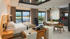 Ein modernes Hotelzimmer mit Bett, Essbereich, Küche und großen Fenstern mit Meerblick.