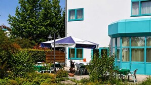 A building with a white exterior and teal accents, outdoor seating with umbrellas, and a garden area.