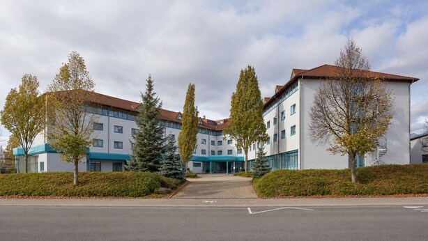 A multi-story hotel with a central entrance, surrounded by trees and a paved driveway.