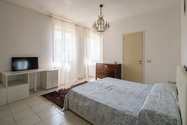 A bedroom with a bed, a television on a stand, a dresser, a door, and a chandelier.