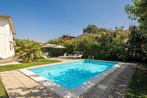 A rectangular swimming pool with a clear blue water surface, surrounded by a gravel border. There are lounge chairs and a table with umbrellas, and a palm tree is visible.