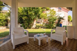 A covered outdoor seating area with two wicker chairs and a small table, surrounded by a checkered floor and greenery.