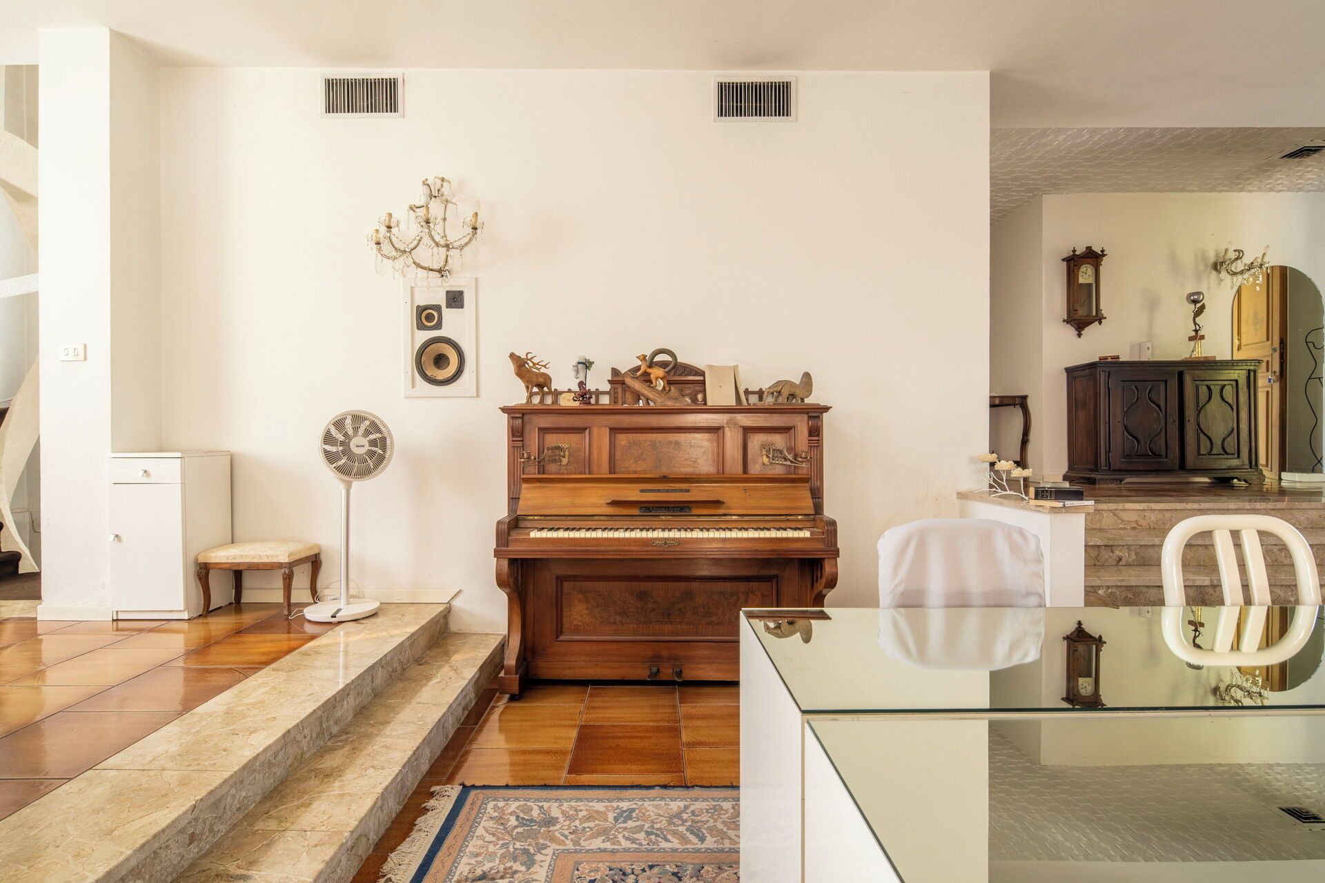 Ein Wohnzimmer mit einem Holzpiano, einem weißen Sessel, einem Glastisch und einem gemusterten Teppich.