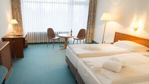 A hotel room with two beds, a desk, two chairs, a lamp, and a view of the outdoors through sheer curtains.