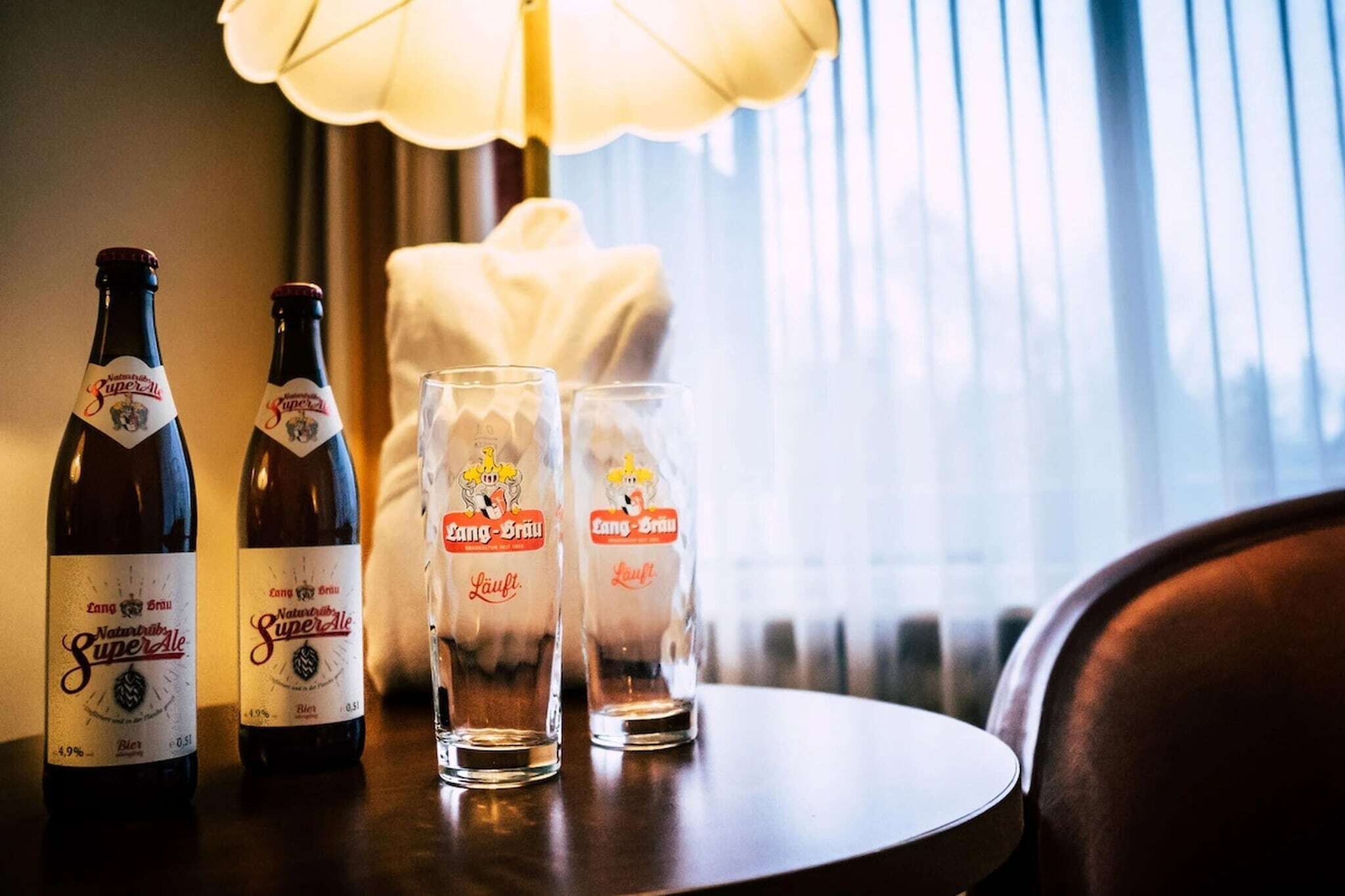 Two bottles of beer and two glasses on a table with a lamp in the background.