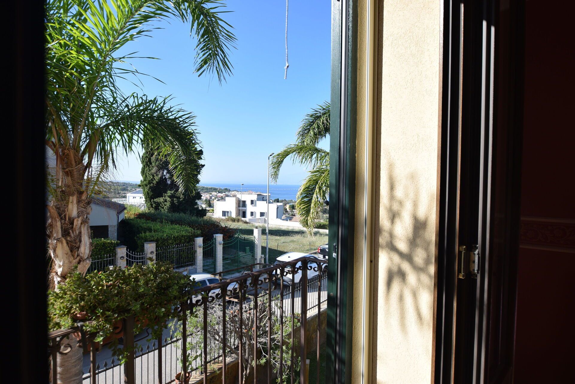 A view from a window overlooking a street with palm trees, a fence, and buildings in the distance.