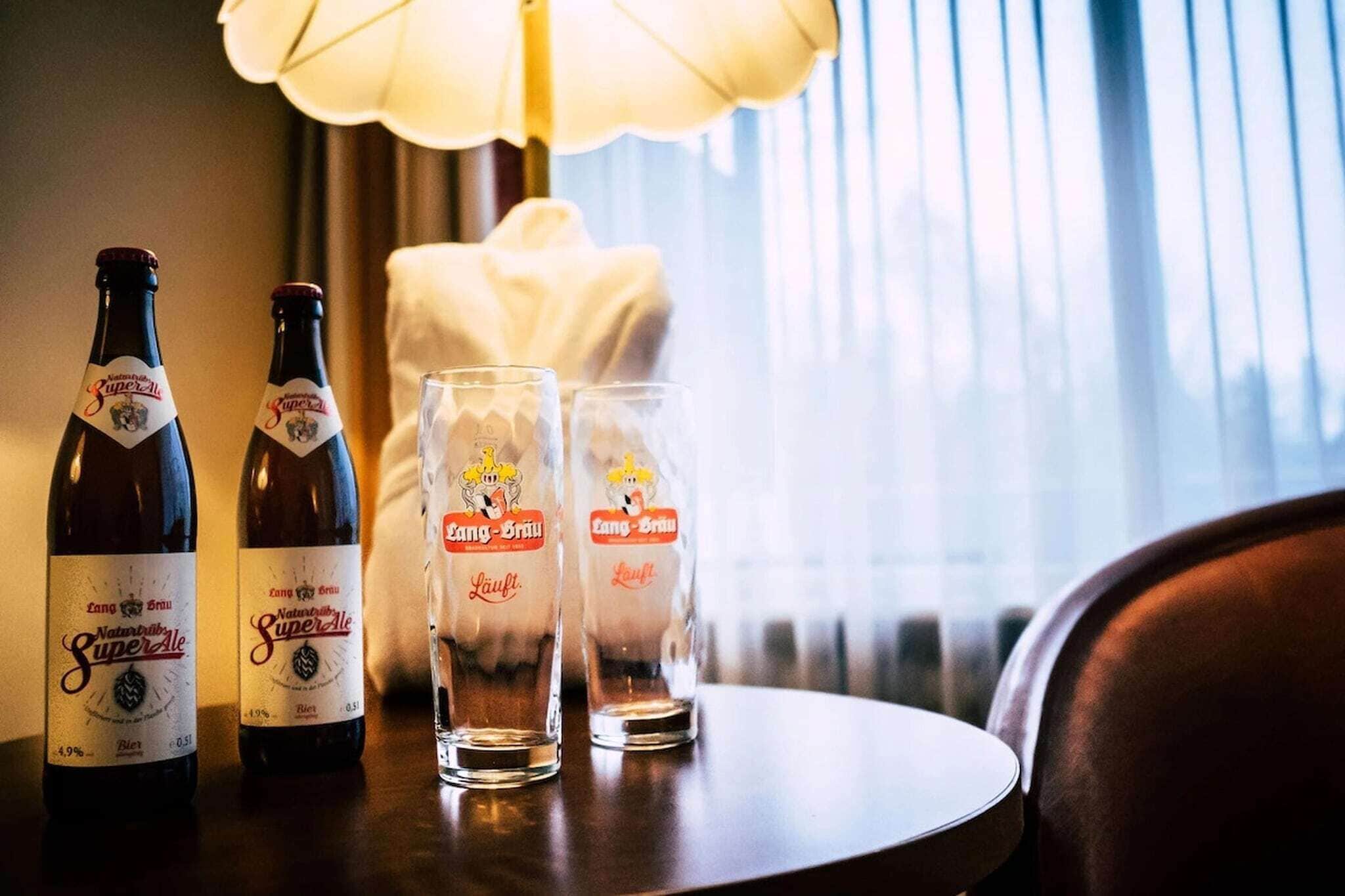 Two bottles of beer and two glasses on a table with a lamp in the background.