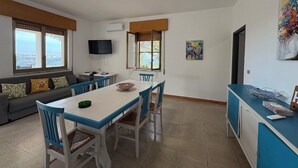 Een zithoek met een tafel en stoelen, een bank met kussens, een aan de muur bevestigde televisie en een raam met uitzicht.