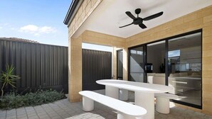 An outdoor patio with a ceiling fan, a white table and bench, and a view into the house.