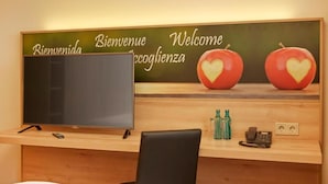 A hotel reception desk with a flat-screen TV, a wooden counter, a black chair, a phone, and two heart-shaped apples on a wall.