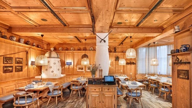 A wooden interior with multiple dining tables and chairs, a central island with a laptop, and hanging light fixtures.