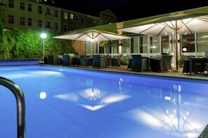 Ein Swimmingpool mit Liegestühlen und Sonnenschirmen, umgeben von einem Gebäude mit großen Fenstern.