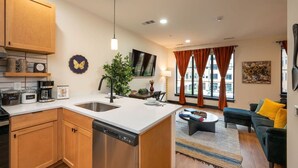 A modern kitchen with a dishwasher, coffee maker, and cabinets. There is a dining area with a sofa and a coffee table.