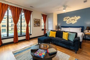 Una sala de estar moderna con un sofá, una mesa de centro con libros, una cama con un letrero de neón y ventanas amplias con cortinas.