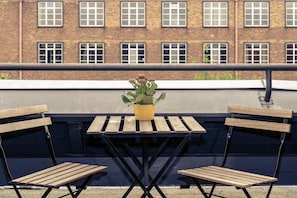 A rooftop terrace with a table and two wooden chairs, a potted plant, and a brick building in the background.