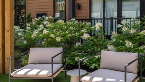 Ein überdachter Außenbereich mit Sitzgelegenheit, zwei Stühlen und einem Tisch, umgeben von blühenden Sträuchern, unter einer Holzpergola.