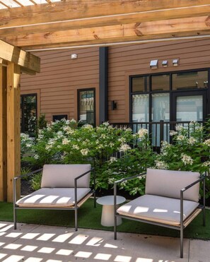 Ein überdachter Außenbereich mit Sitzgelegenheit, zwei Stühlen und einem Tisch, umgeben von blühenden Sträuchern, unter einer Holzpergola.