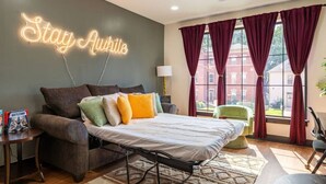 Ein Zimmer mit Bett, Sofa, einer Neonreklame und großen Fenstern mit Vorhängen.