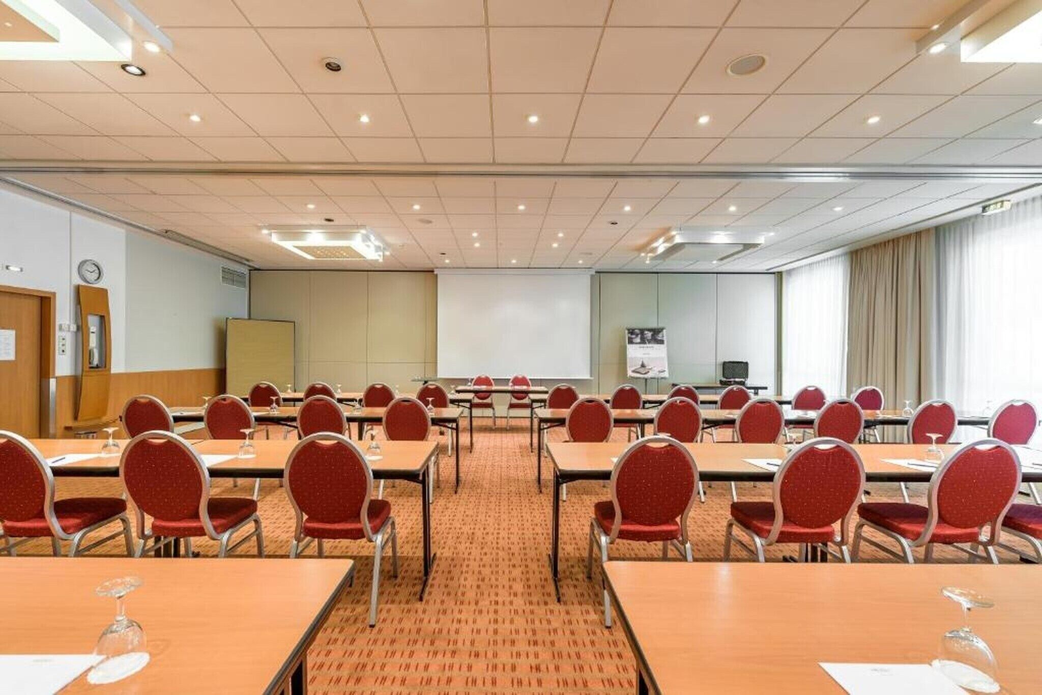 Uma sala de conferências com várias mesas e cadeiras, uma tela de projeção e um relógio na parede.