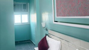 Une chambre d’hôtel aux murs de couleur turquoise, un lit avec des oreillers blancs et bordeaux, une grande fenêtre et une décoration murale à motifs.