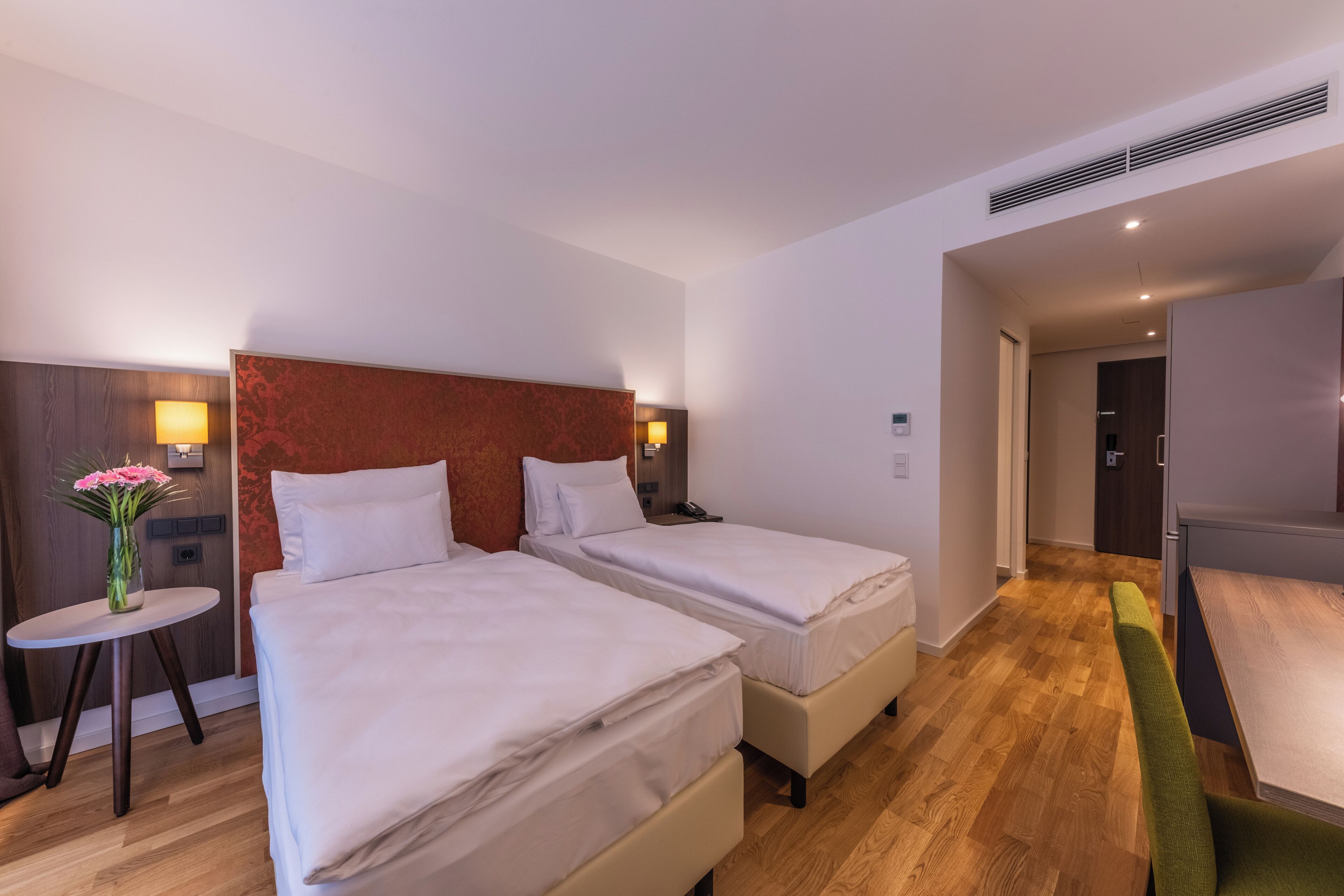 Ein Hotelzimmer mit zwei Betten, einem kleinen Tisch mit Blumenvase, einem Schreibtisch mit Lampe und Holzboden.