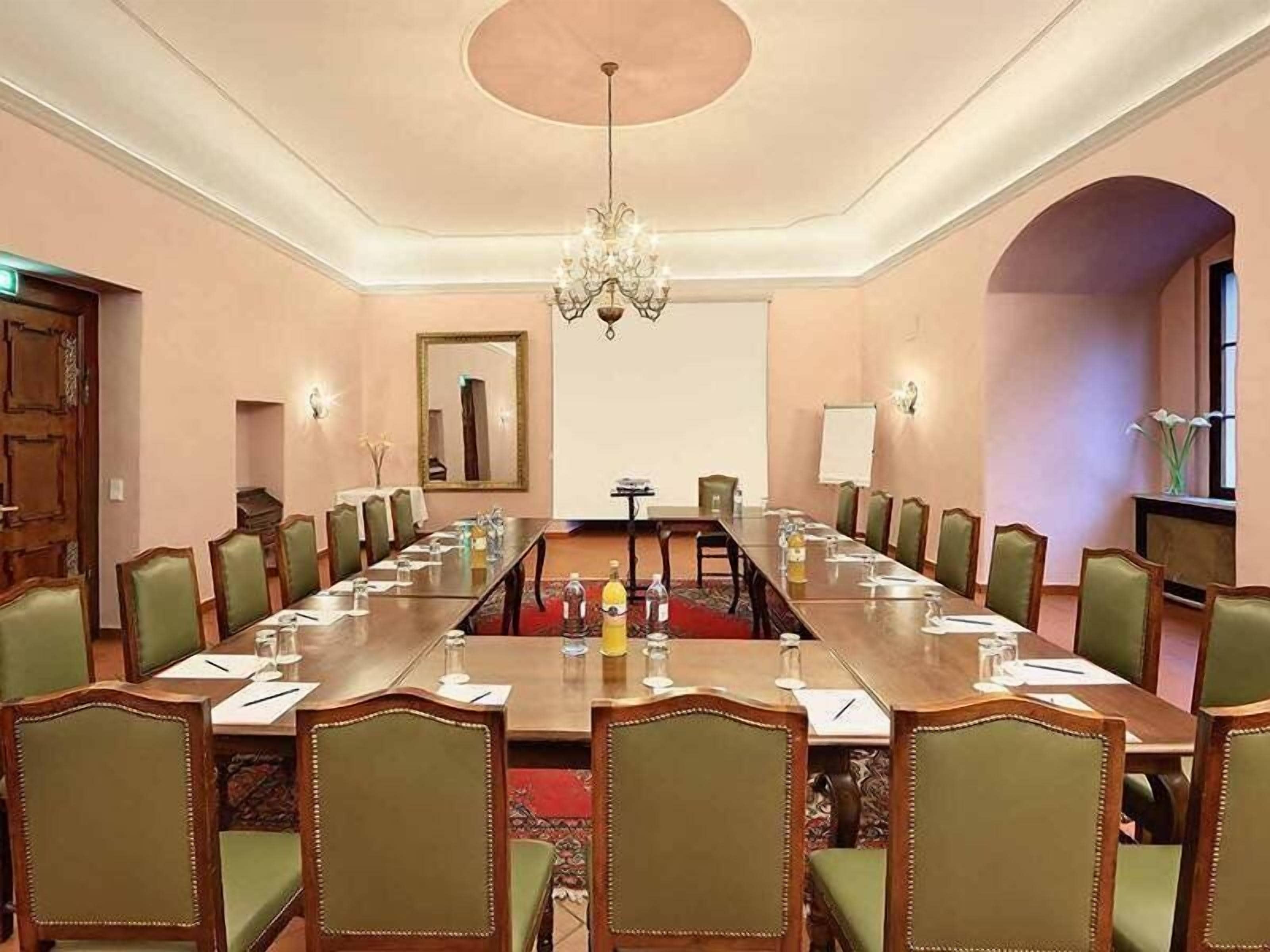 Un salón de conferencias con una larga mesa, sillas tapizadas en verde, una araña y un espejo en la pared.
