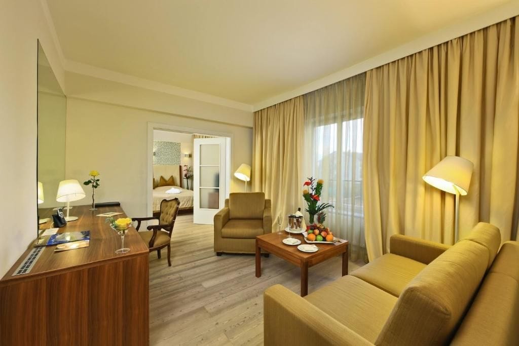 Ein Hotelzimmer mit einem hölzernen Schreibtisch, einem Sofa, einem Sessel, einem Bett und einem großen Fenster mit Vorhängen.