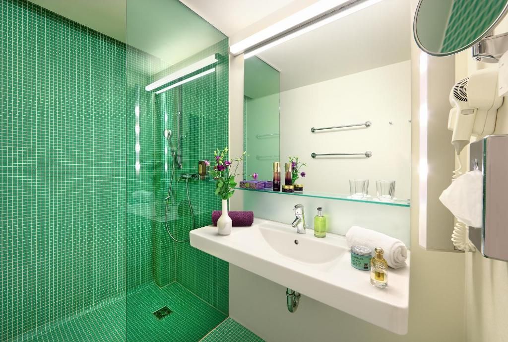 A modern bathroom with a green-tiled shower, a white sink with toiletries, and a mirror with a towel rack.