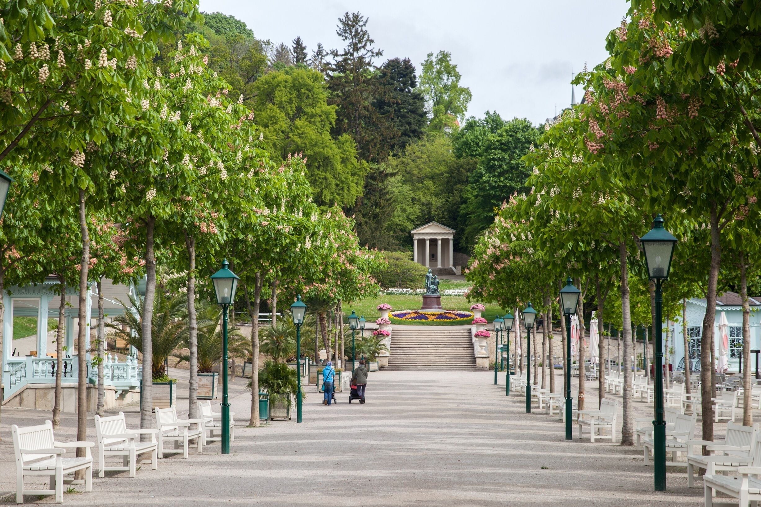 A park pathway with benches and lampposts, leading to a statue and steps.