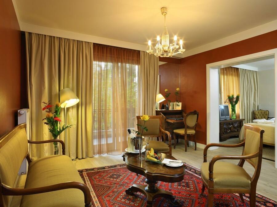 Ein Hotelzimmer mit Essbereich, einer Kandelaberleuchte, einem großen Fenster mit Vorhängen und einem Spiegel, der den Raum widerspiegelt.