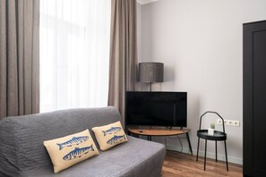 Ein modernes Wohnzimmer mit grauem Sofa, zwei Kissen mit Fischmuster, einem Flachbildfernseher auf einem Holzbodenständchen, einem kleinen Beistelltisch und einem schwarzen Sessel.
