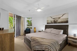 Ein Schlafzimmer mit einem großen Bett, einem Deckenventilator, einem Nachttisch, einer Lampe und einem Fenster mit Blick auf Grünflächen.