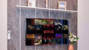 A wall-mounted TV with a Netflix logo, surrounded by a marble-patterned wall, a white shelf with decorative items, and a potted plant. - Corporate Stays Zuba 8 -116 (Fernando de la Mora)